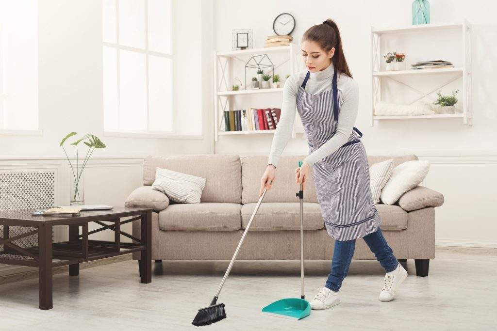 Efficient professional maid sweeping living room floor with broom and dustpan.
